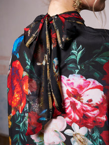 The image features the back of a person in the Social Queen Artistic Floral Print Ruffle Dress, a chic black dress with red, pink, and white blooms and a statement neck bow—ideal for a stylish wedding guest look.