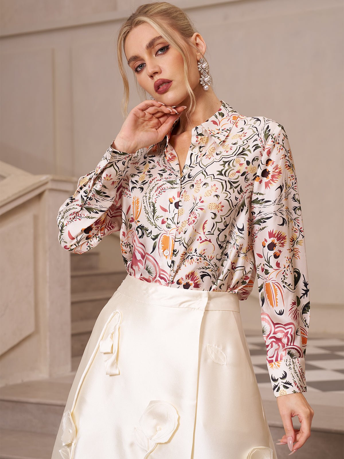 A woman stands indoors near a staircase, looking at the camera, dressed in the Social Queen Artistic Floral Print Silk Blouse and a cream satin skirt with a pearlescent sheen.