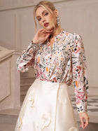 A woman stands indoors near a staircase, looking at the camera, dressed in the Social Queen Artistic Floral Print Silk Blouse and a cream satin skirt with a pearlescent sheen.