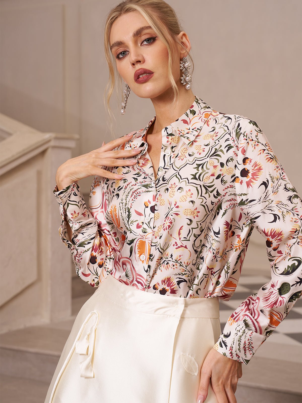 A woman stands indoors, wearing the Social Queen Artistic Floral Print Silk Blouse with long sleeves and a cream skirt, posing with one hand on her chest and the other on her hip.