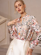 A woman stands indoors, wearing the Social Queen Artistic Floral Print Silk Blouse with long sleeves and a cream skirt, posing with one hand on her chest and the other on her hip.
