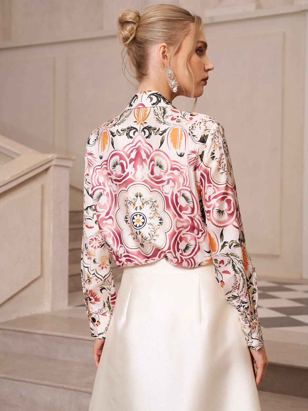 A woman with blonde hair in an updo wears the Social Queen Artistic Floral Print Silk Blouse and a white skirt, standing on stairs with her back mostly to the camera.