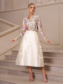 A woman stands on stairs wearing the Social Queen Artistic Floral Print Silk Blouse and a cream midi skirt with flower appliqués, paired with heeled sandals.