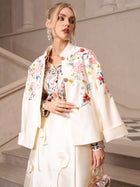 A woman stands indoors near a staircase, her outfit catching the light as she wears the Social Queen Artistic Floral Print Silk Blouse paired with a matching cream skirt and jacket.