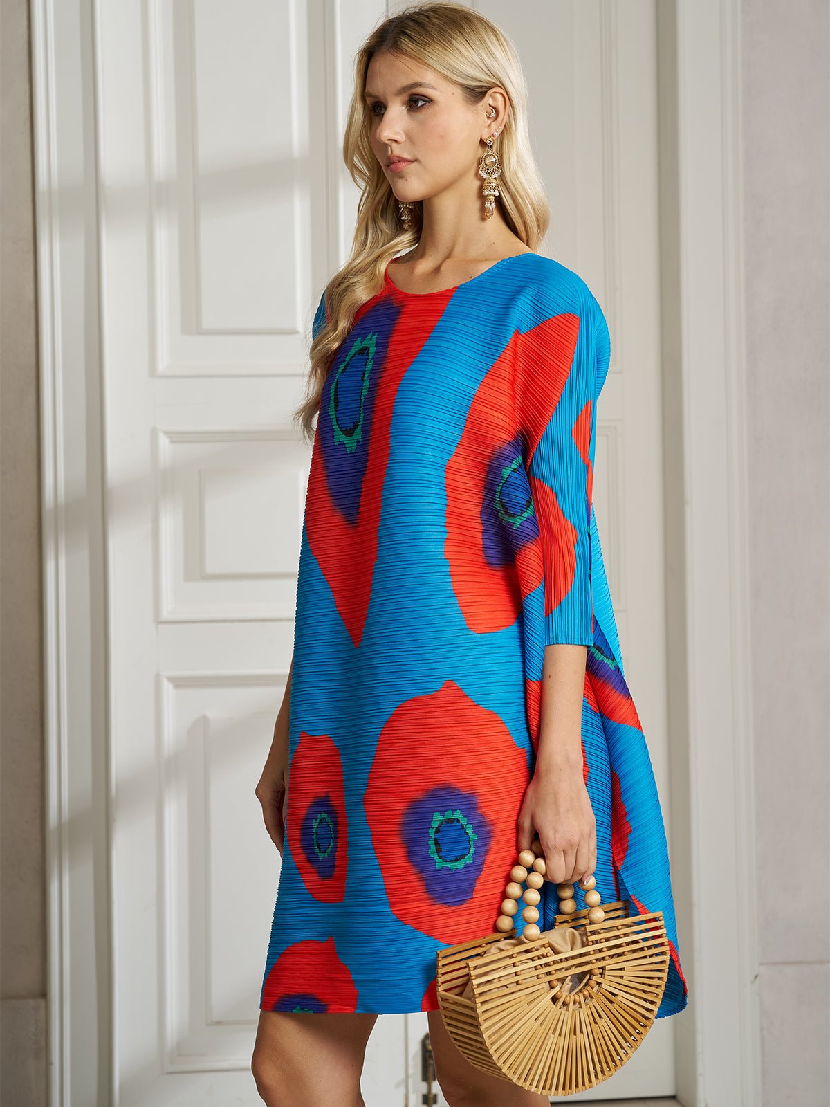 A woman with long blonde hair and large earrings stands indoors holding a wooden handbag, wearing the Social Queen Artistic Print Plissé A-Line Dress for a chic resort-ready look.