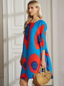 A woman with long blonde hair and large earrings stands indoors holding a wooden handbag, wearing the Social Queen Artistic Print Plissé A-Line Dress for a chic resort-ready look.