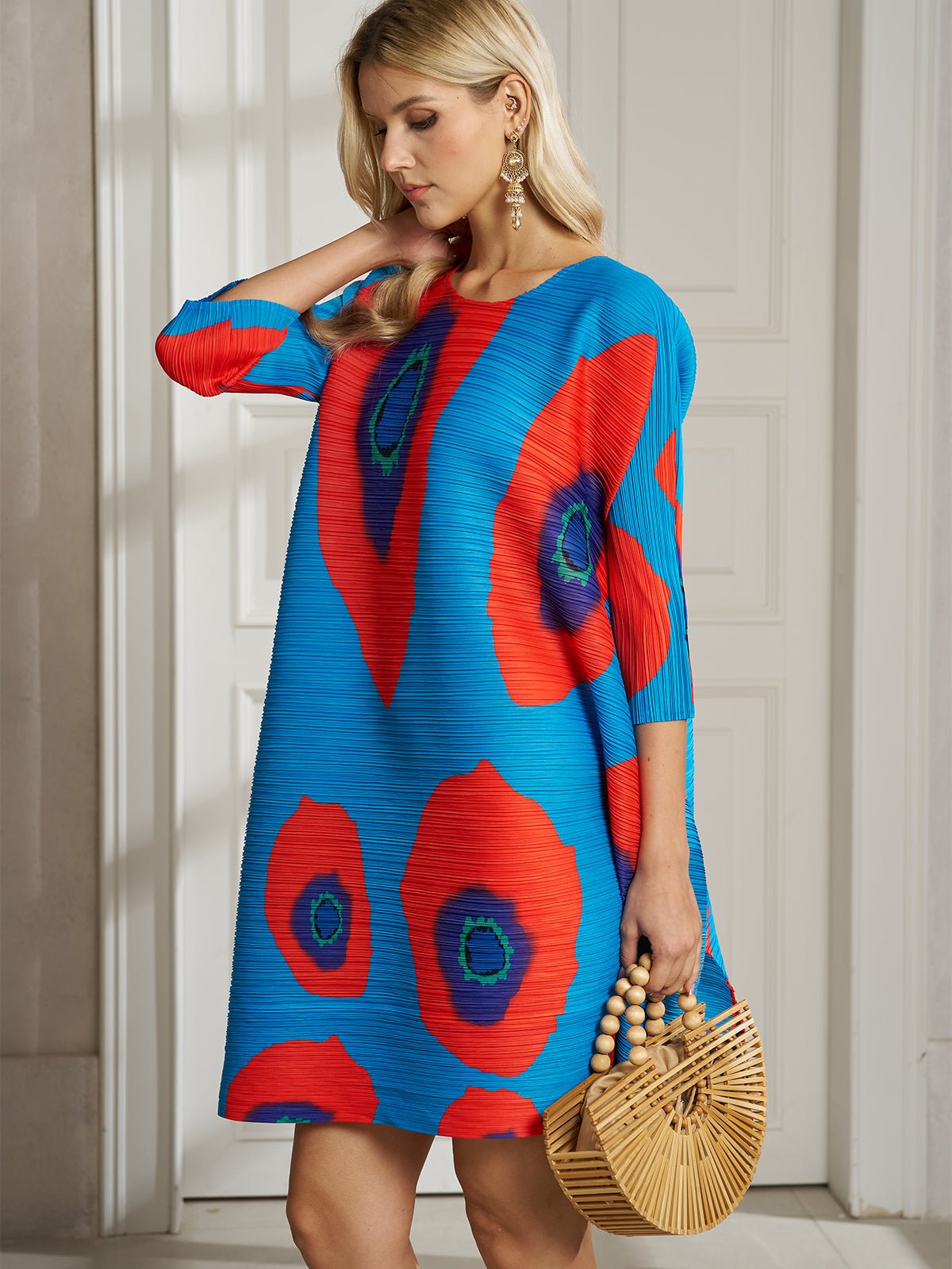 A woman in the Social Queen Artistic Print Plissé A-Line Dress with bold red and green circles holds a bamboo handbag while standing indoors near a white door.