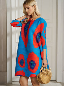 A woman in the Social Queen Artistic Print Plissé A-Line Dress with bold red and green circles holds a bamboo handbag while standing indoors near a white door.