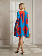 A woman stands indoors facing away, wearing the Artistic Print Plissé A-Line Dress by Social Queen in blue and red with tan heels and a wicker handbag. White double doors are visible in the background.