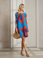 A woman stands indoors wearing the Social Queen Artistic Print Plissé A-Line Dress in blue and red, paired with a wooden handbag and heeled sandals.
