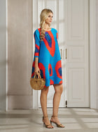 A woman wears the Social Queen Artistic Print Plissé A-Line Dress indoors, paired with a wooden handbag and gold heeled sandals.