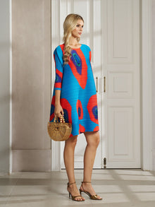 A woman wears the Social Queen Artistic Print Plissé A-Line Dress indoors, paired with a wooden handbag and gold heeled sandals.