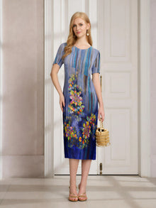 A woman indoors wears the Social Queen Artistic Smudge Print Plissé Dress, featuring blue ombre floral stripes and short sleeves—styled with sandals and a woven handbag for ideal resort wear.