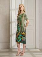 A woman stands sideways in front of a white paneled door wearing the Social Queen Artistic Smudge Print Plissé Dress in green, styled with gold high-heeled sandals.