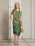A woman stands indoors in the Social Queen Artistic Smudge Print Plissé Dress, a knee-length, short-sleeve pleated piece with vibrant ombre stripes and florals, paired with a gold bag and sandals.