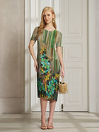 A woman stands indoors in the Social Queen Artistic Smudge Print Plissé Dress, a knee-length, short-sleeve pleated piece with vibrant ombre stripes and florals, paired with a gold bag and sandals.
