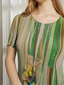 A woman wears the Artistic Smudge Print Plissé Dress by Social Queen—a short-sleeved silk style with vertical green, yellow, and beige stripes, plus a floral accent at the waist—ideal for effortless resort wear.