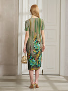 A woman with long red hair stands by a white door, wearing Social Queen's Artistic Smudge Print Plissé Dress and holding a small gold handbag—an elegant choice for chic resort style.