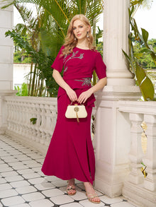 A woman stands by a white balustrade, holding a white purse, wearing the Social Queen Asymmetric Beaded Mermaid Slit Dress, with greenery and palm leaves in the background.