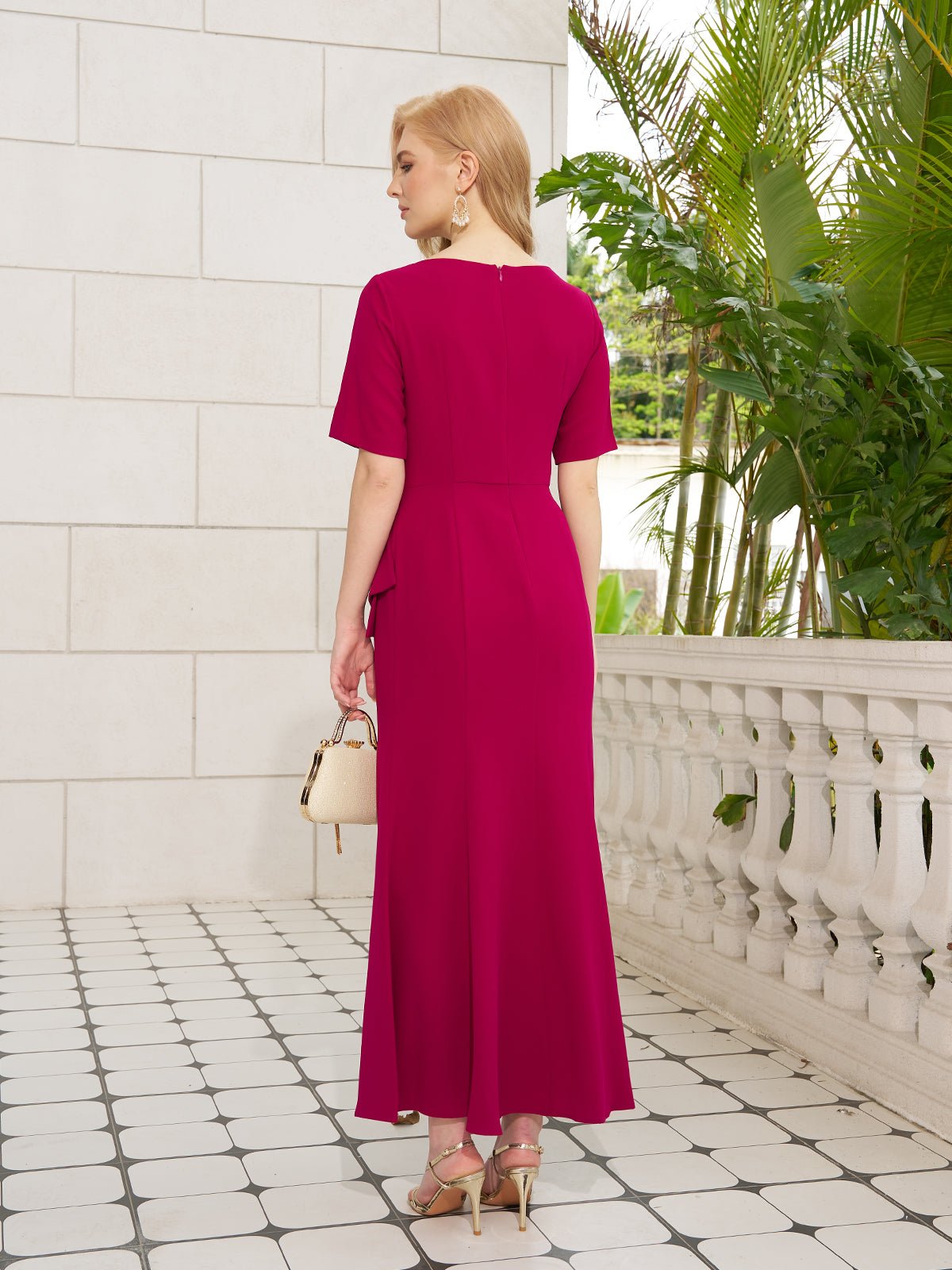 A woman stands on a tiled terrace surrounded by green plants, facing away, wearing the Social Queen Asymmetric Beaded Mermaid Slit Dress in magenta, gold heels, large earrings, and holding a small white handbag.
