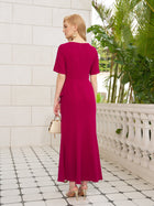 A woman stands on a tiled terrace surrounded by green plants, facing away, wearing the Social Queen Asymmetric Beaded Mermaid Slit Dress in magenta, gold heels, large earrings, and holding a small white handbag.
