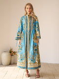 A woman stands indoors in the Social Queen Baroque Engineered-Print Cotton Dress, a long maxi silhouette with blue and gold Baroque floral design, beside a vase of flowers on a tiled floor.