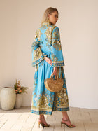 A woman in the Social Queen Baroque Engineered-Print Cotton Dress, featuring a blue and gold floral motif and maxi silhouette, stands sideways with a bamboo handbag and heeled sandals; vases and flowers are visible behind her.