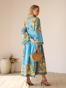A woman in the Social Queen Baroque Engineered-Print Cotton Dress, featuring a blue and gold floral motif and maxi silhouette, stands sideways with a bamboo handbag and heeled sandals; vases and flowers are visible behind her.