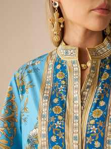 A woman wears the Social Queen Baroque Engineered-Print Cotton Dress, featuring blue and gold floral patterns with a mandarin collar, paired with large gold earrings; only her lower face and upper torso are shown.