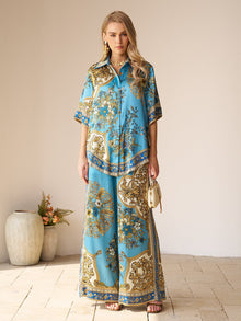 A woman stands indoors wearing the Social Queen Baroque Engineered-Print Silk Shirt and matching wide-leg pants, holding a small cream handbag. Decorative vases with flowers are in the background.
