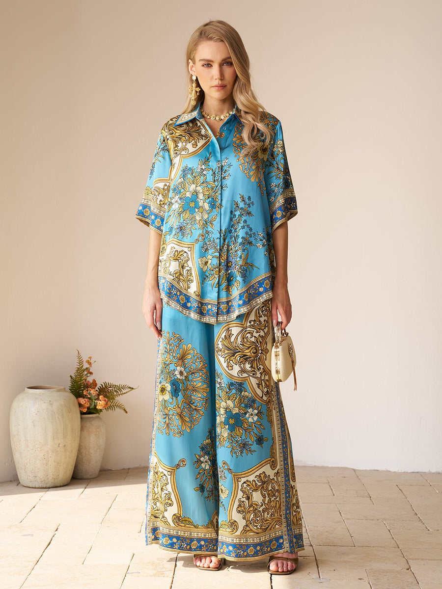 A woman stands indoors wearing the Social Queen Baroque Engineered-Print Silk Shirt and matching wide-leg pants, holding a small cream handbag. Decorative vases with flowers are in the background.