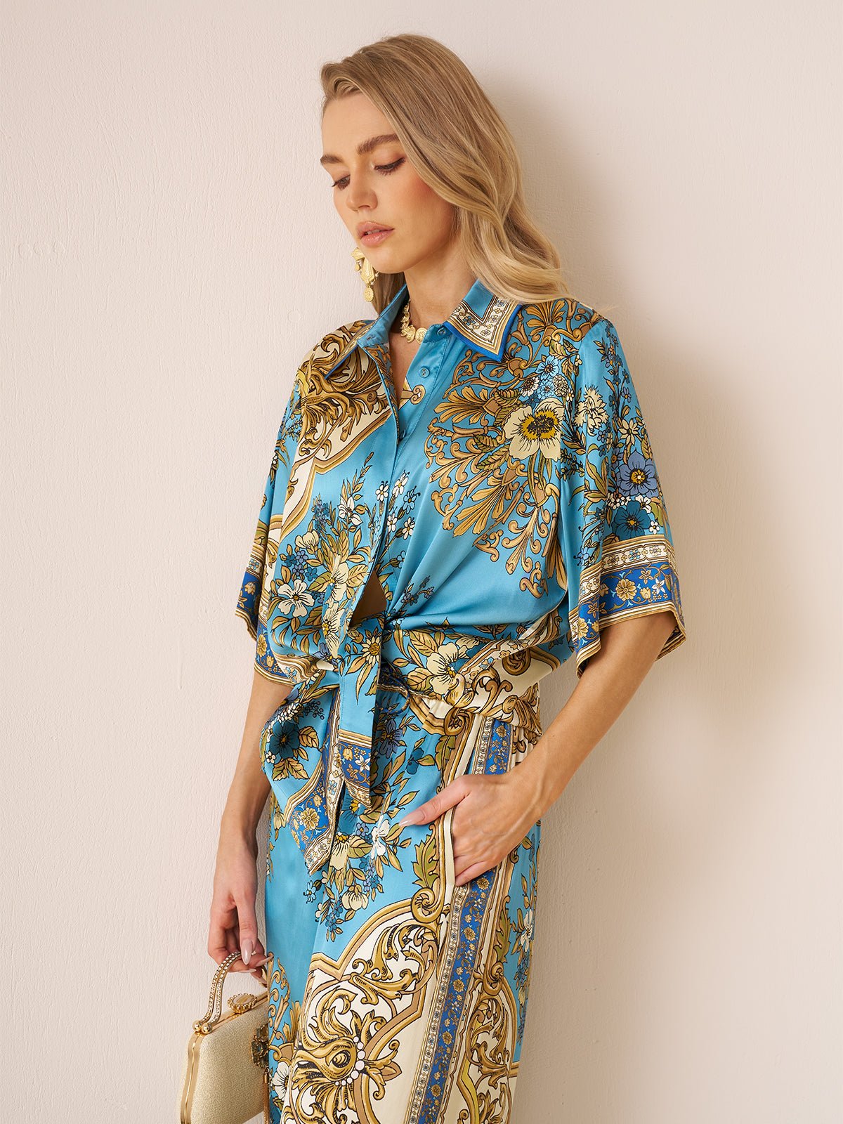 A woman stands against a light wall, wearing the Social Queen Baroque Engineered-Print Silk Shirt with blue and gold floral designs, and holding a small gold handbag.