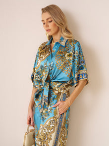 A woman stands against a light wall, wearing the Social Queen Baroque Engineered-Print Silk Shirt with blue and gold floral designs, and holding a small gold handbag.