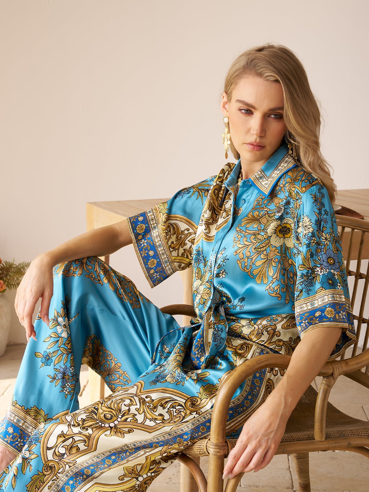 A woman sits on a wooden chair wearing the Social Queen Baroque Engineered-Print Silk Shirt in blue and gold, her arm resting on the chair’s back, gazing ahead with a neutral expression.