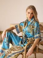 A woman sits on a wooden chair wearing the Social Queen Baroque Engineered-Print Silk Shirt in blue and gold, her arm resting on the chair’s back, gazing ahead with a neutral expression.