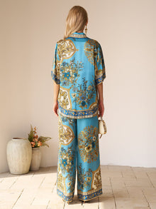 A woman stands indoors, facing away, wearing the Social Queen Baroque Engineered-Print Silk Shirt with matching wide-leg pants and holding a small white handbag. Decorative vases with flowers are nearby.