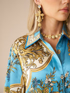 Woman shown from the shoulder up wearing the Social Queen Baroque Engineered-Print Silk Shirt, accessorized with a gold medallion necklace and ornate gold cross-shaped earrings.