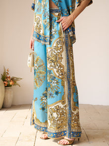 A person wears Social Queen's Baroque Engineered-Print Silk Wide-Leg Pants with a matching top, holding a small beige bag, standing on a tiled floor near a potted plant.