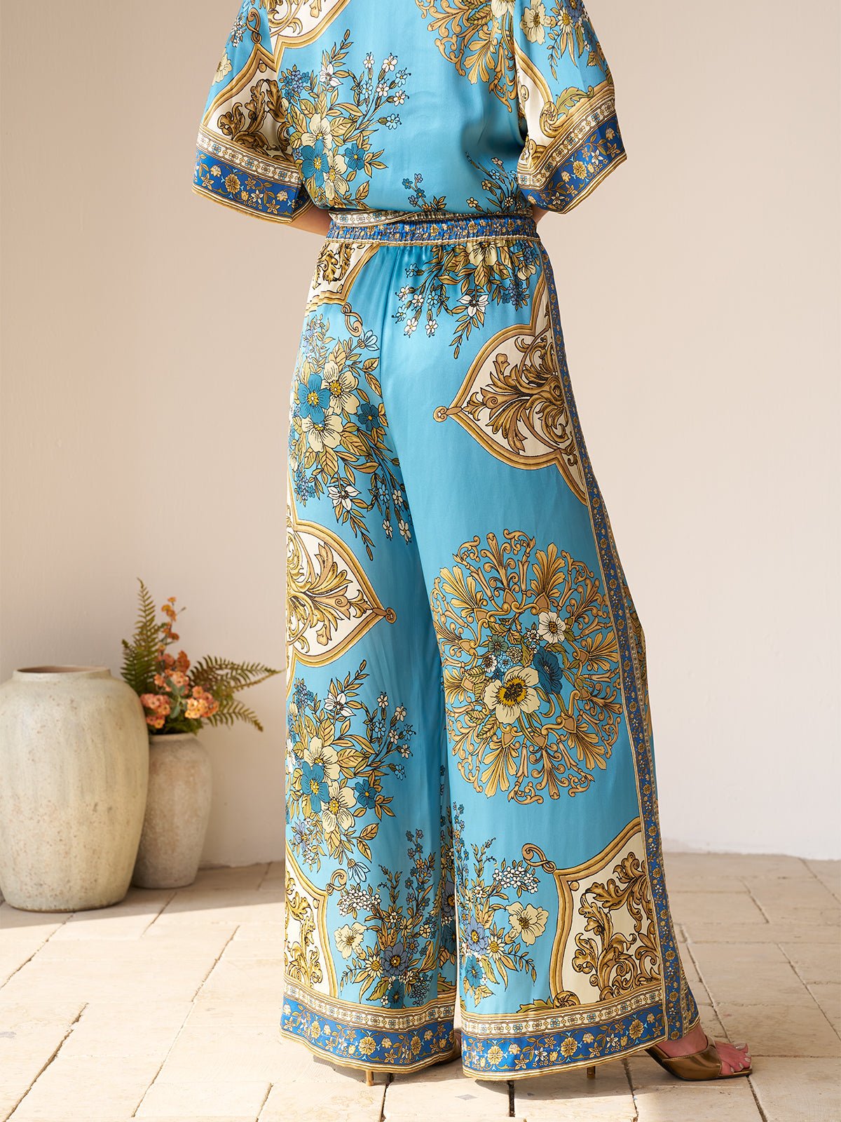 A person stands indoors with their back to the camera, wearing Social Queen’s Baroque Engineered-Print Silk Wide-Leg Pants and a matching top. Decorative vases and greenery appear in the background.