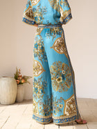 A person stands indoors with their back to the camera, wearing Social Queen’s Baroque Engineered-Print Silk Wide-Leg Pants and a matching top. Decorative vases and greenery appear in the background.