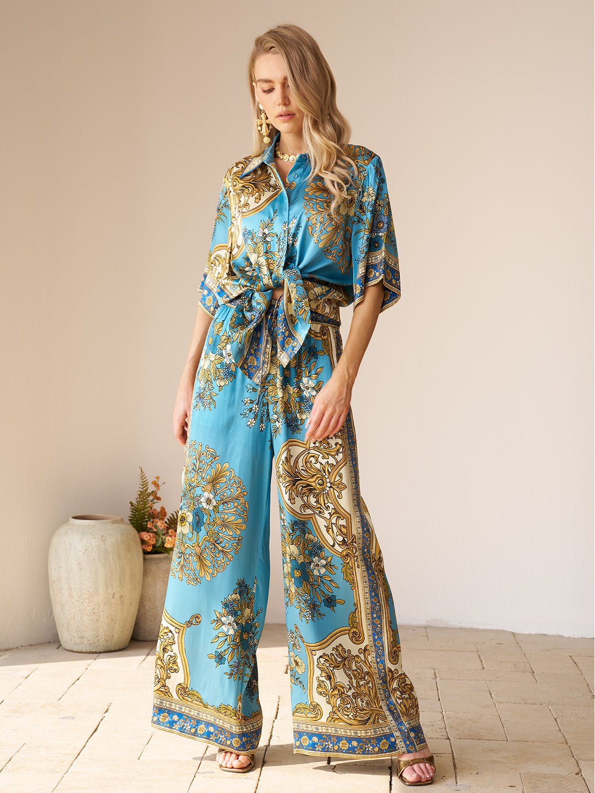 A woman stands indoors wearing Social Queen's Baroque Engineered-Print Silk Wide-Leg Pants with a tied waist. A ceramic vase with flowers sits on the floor beside her.