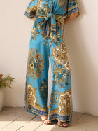 Only the lower half of a person is visible, wearing Social Queen Baroque Engineered-Print Silk Wide-Leg Pants in blue and gold, belted at the waist and styled with gold sandals, standing against a wall.