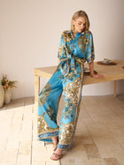 A woman wearing Social Queen’s Baroque Engineered-Print Silk Wide-Leg Pants leans against a wooden table in a minimalist room with tiled flooring and potted plants.