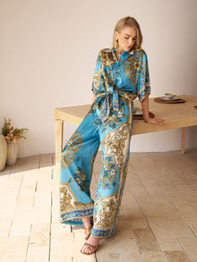 A woman wearing Social Queen’s Baroque Engineered-Print Silk Wide-Leg Pants leans against a wooden table in a minimalist room with tiled flooring and potted plants.