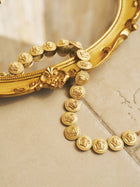 The Social Queen Baroque Medallion Necklace is draped over the edge of an ornate framed mirror, resting on a beige tiled surface.