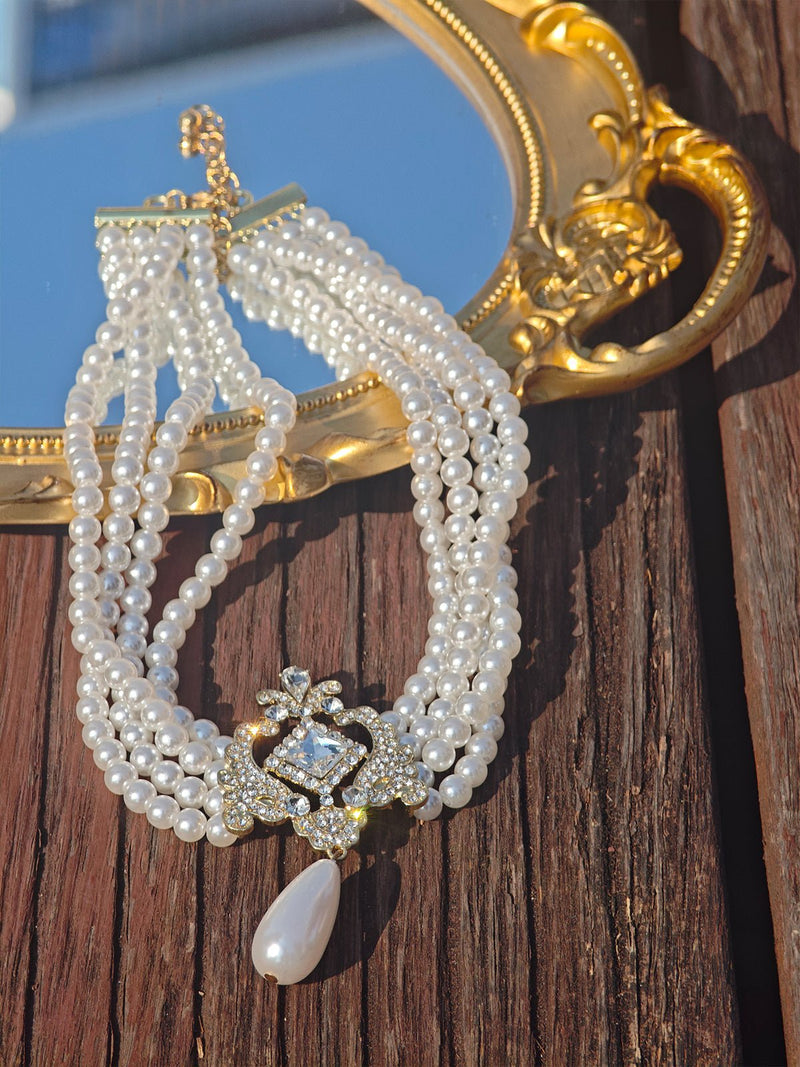 The Social Queen Baroque Pearl Layered Necklace, featuring gold-tone crystal accents and a jeweled teardrop pendant, is displayed on a wooden surface beside a gold-framed mirror.