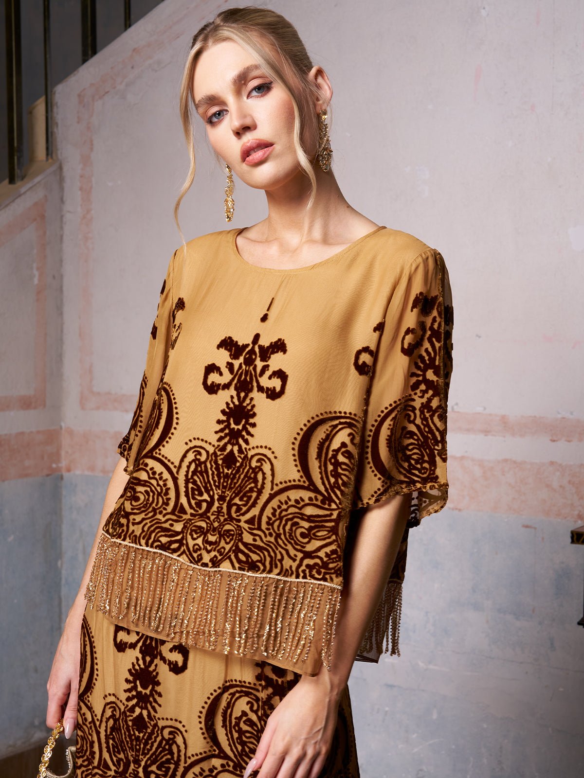 A woman models the Social Queen Baroque Velvet Fringe Set, a tan silk burnout velvet outfit with dark baroque patterns and fringe details, standing before a textured painted wall.