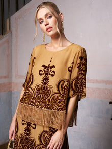 A woman models the Social Queen Baroque Velvet Fringe Set, a tan silk burnout velvet outfit with dark baroque patterns and fringe details, standing before a textured painted wall.