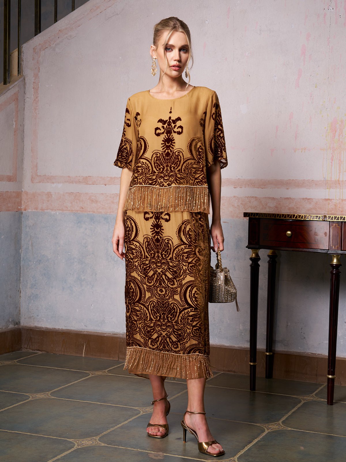 A woman stands indoors in the Social Queen Baroque Velvet Fringe Set—gold and brown silk burnout velvet with baroque patterns and fringe—paired with a handbag and gold heels for an elegant wedding guest look.