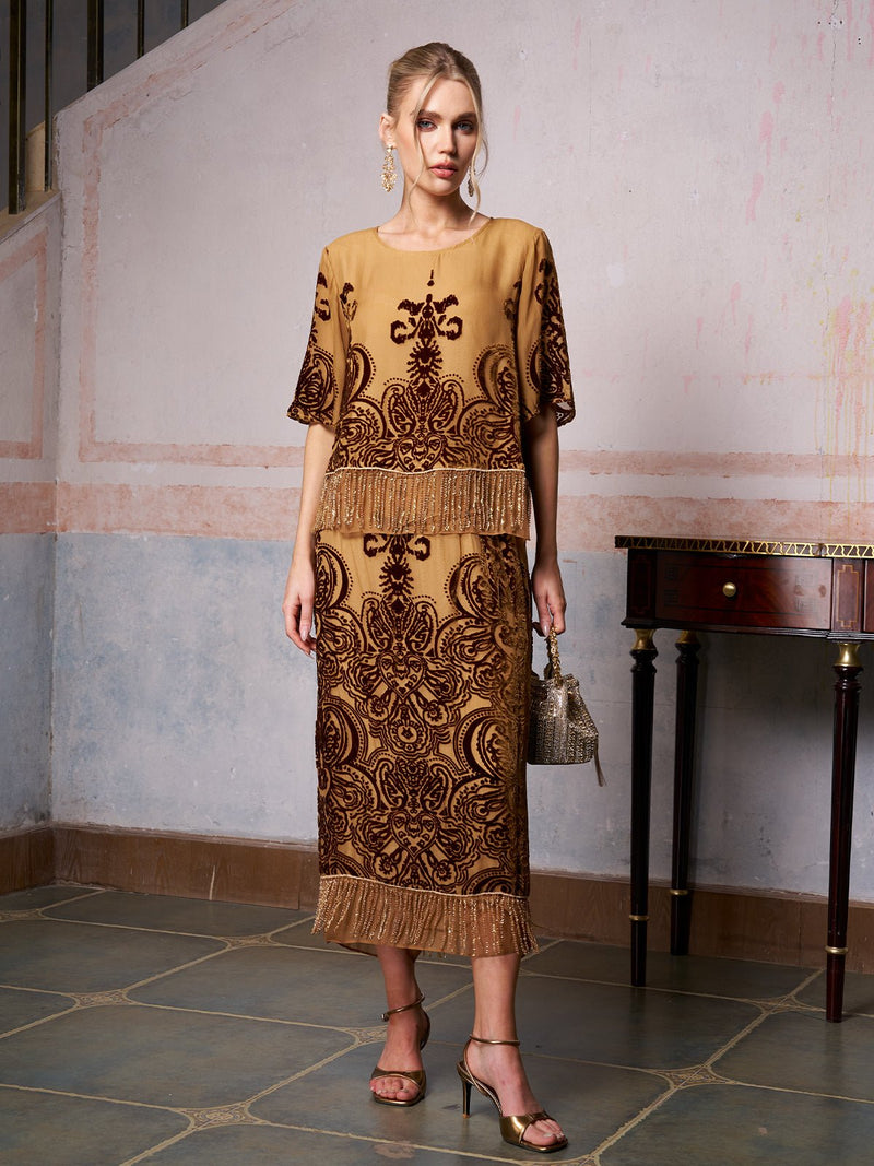 A woman poses indoors in the Baroque Velvet Fringe Set by Social Queen—a gold and brown silk burnout velvet outfit with fringe details. Paired with gold heels and a small handbag, it’s a chic pick for wedding guests.